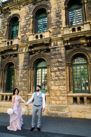 Couple in formal wear standing by historic stone building with green windows.