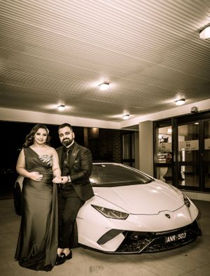 A couple poses beside a white Lamborghini sports car under indoor lighting.