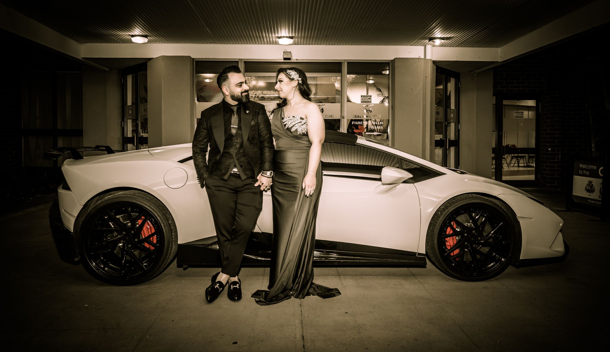 A stylish couple poses next to a white luxury sports car in an evening setting.
