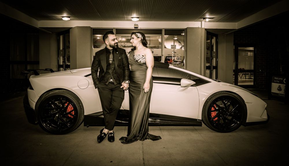A stylish couple poses next to a white luxury sports car in an evening setting.