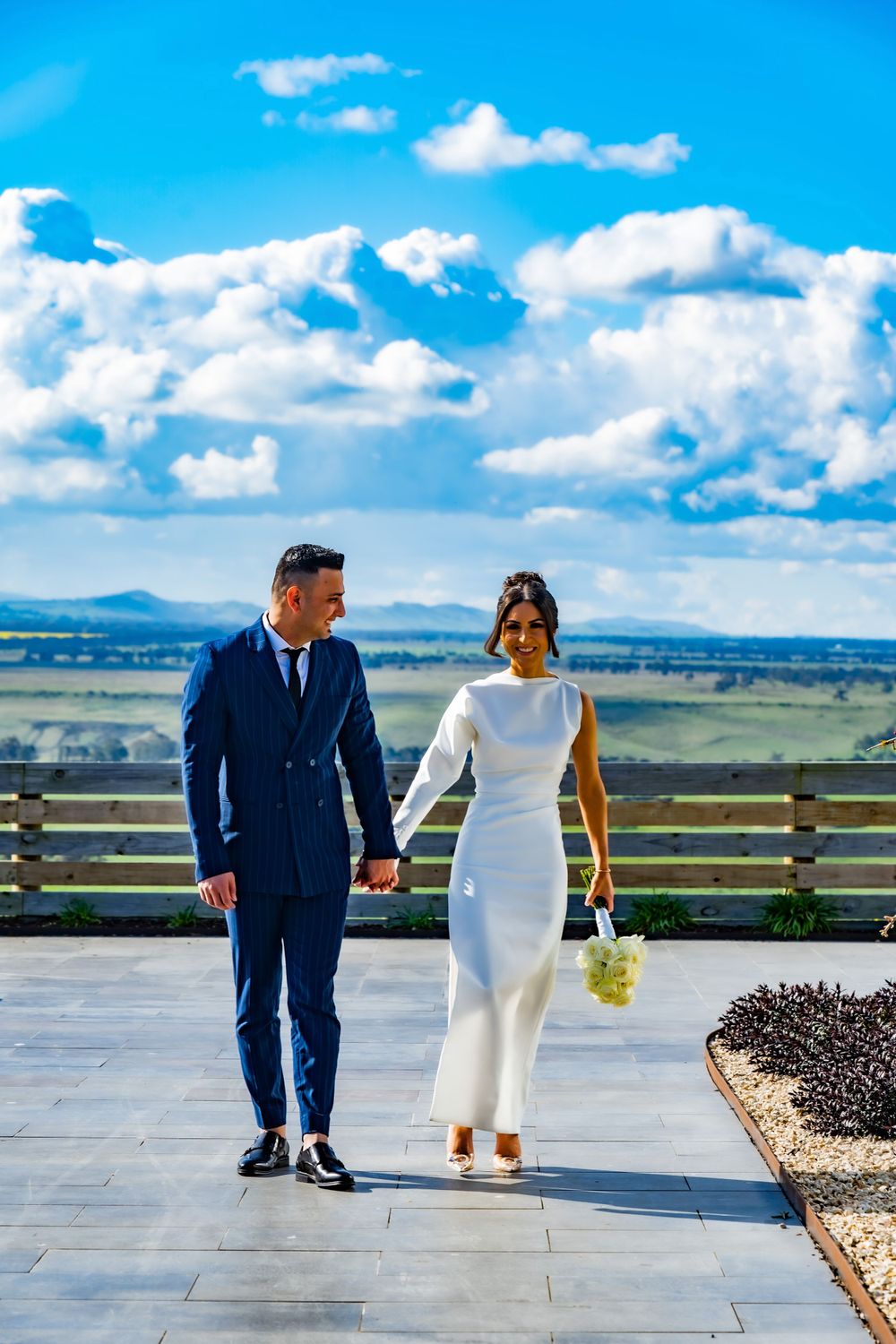Couple in wedding attire walking on terrace with mountain landscape and blue sky.