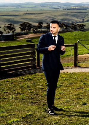 A man in a pinstripe suit adjusts his jacket while standing on a scenic hilltop.