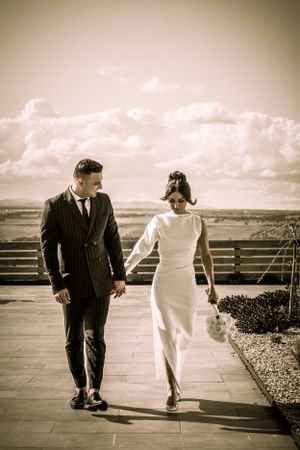Sepia-toned wedding portrait of couple walking on outdoor terrace with cloudy sky.