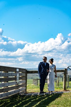 A couple in wedding attire stands by wooden fence under bright blue skies with puffy clouds.