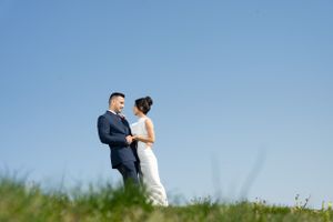 A couple stands together on grassy hilltop against bright blue sky.