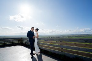 A couple stands together on a scenic overlook with rolling hills and bright blue skies in the background.