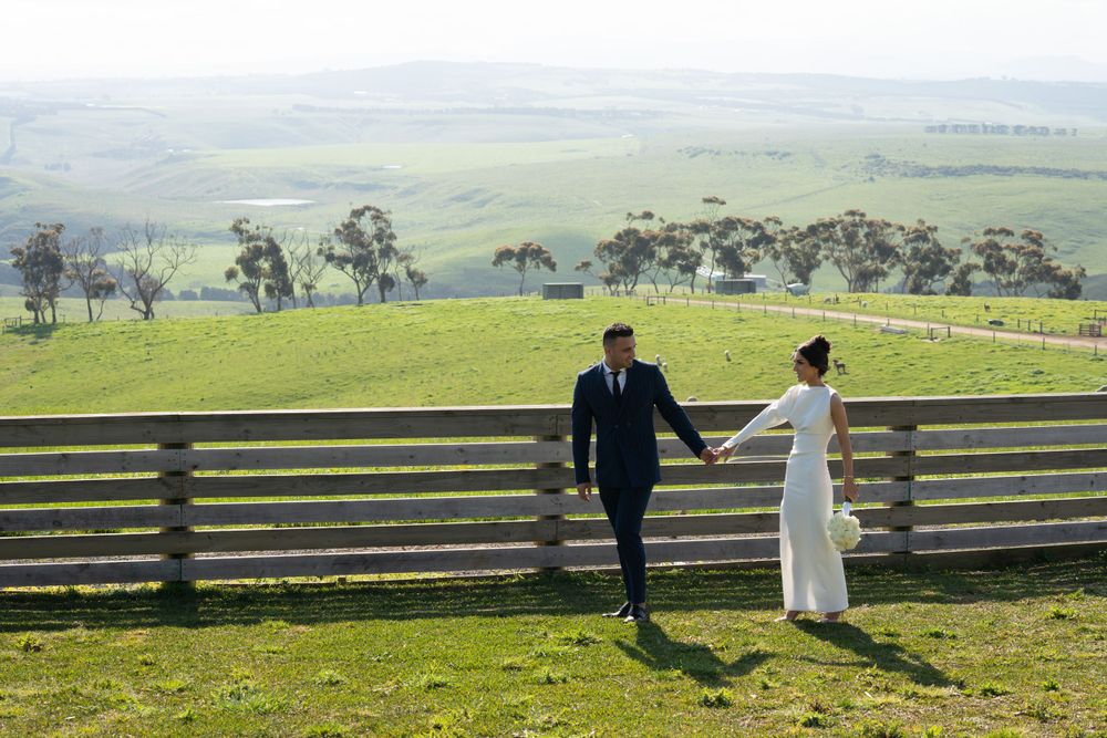 A couple in wedding attire walks hand in hand along a wooden fence overlooking green rolling hills.