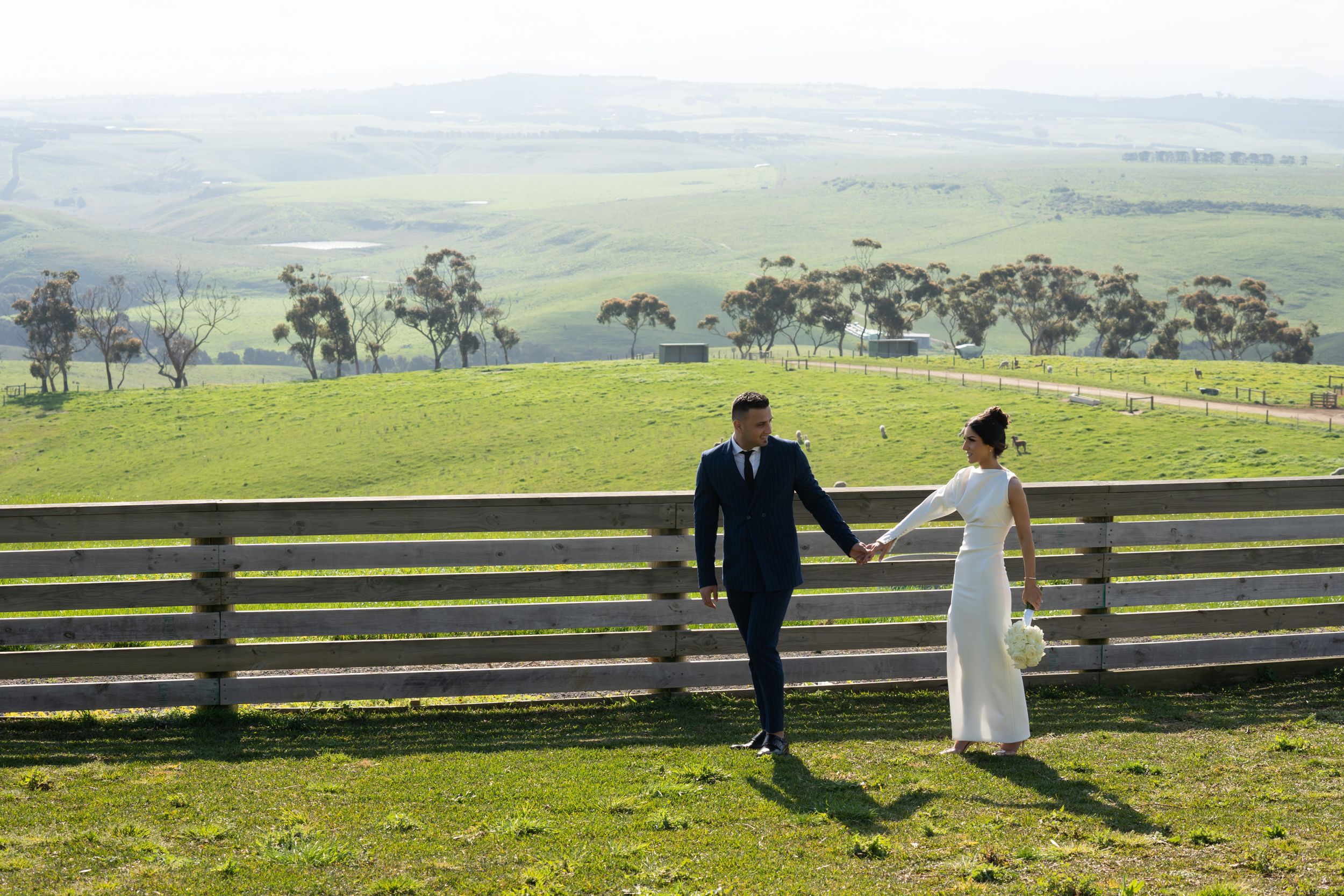 A couple in wedding attire walks hand in hand along a wooden fence overlooking green rolling hills.