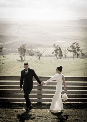 A bride and groom hold hands while walking along a rustic wooden fence in a dreamy countryside setting.
