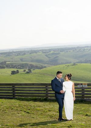 A newlywed couple poses together against a rustic fence with sweeping countryside views.