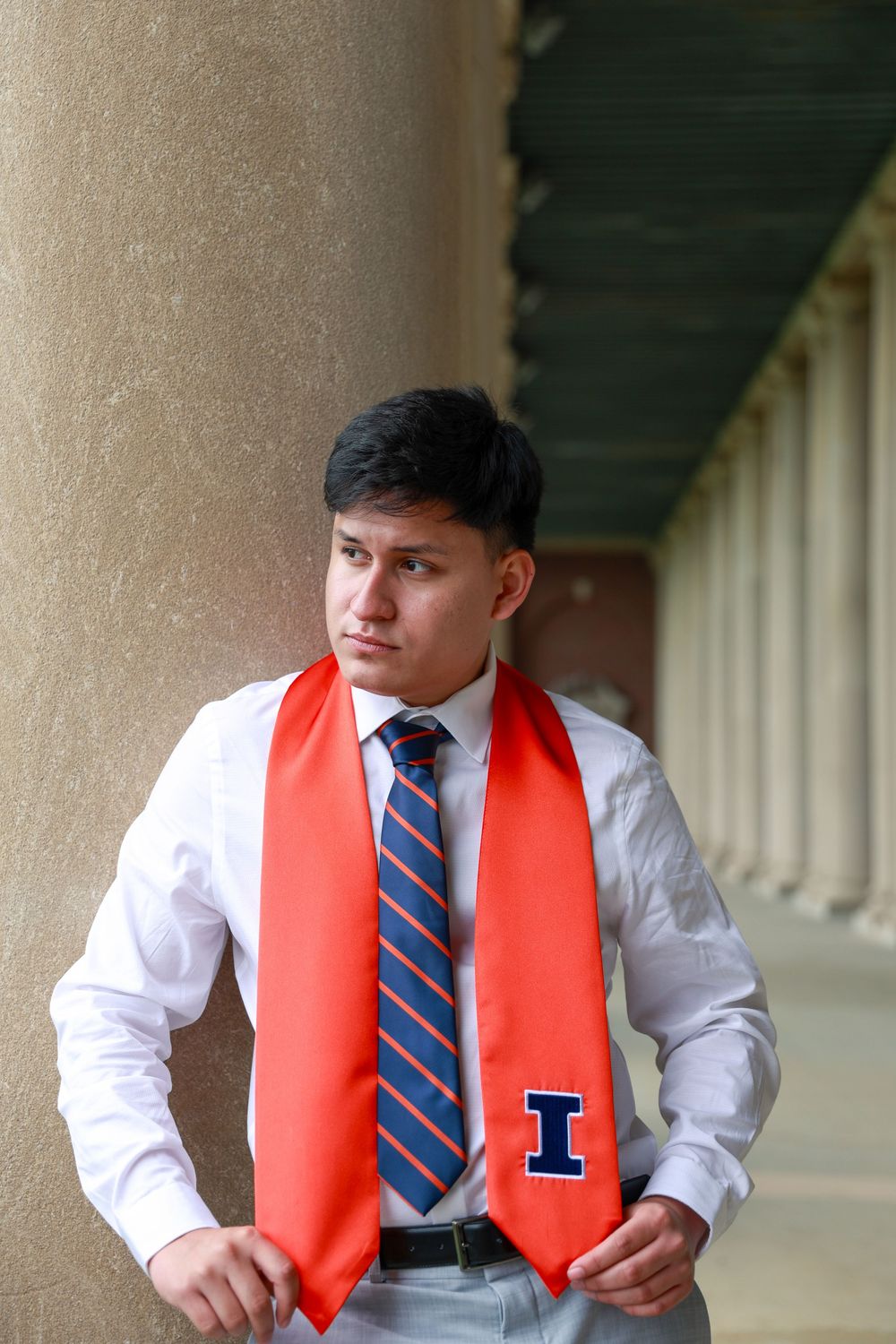 Graduate wearing white shirt and red stole poses against architectural columns.