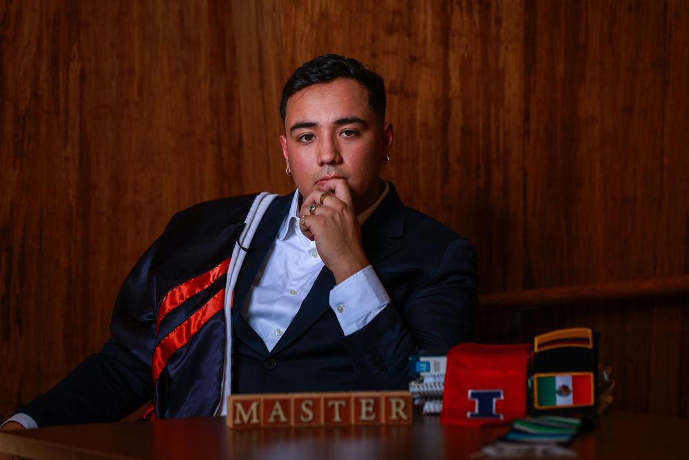 A student in academic regalia sits in contemplation at a desk with wooden paneling in the background.