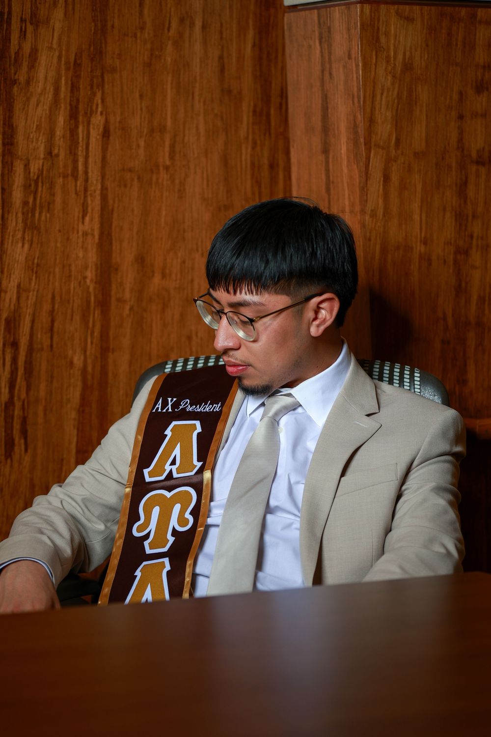 Graduate wearing tan suit and academic stole sits thoughtfully at wooden desk.
