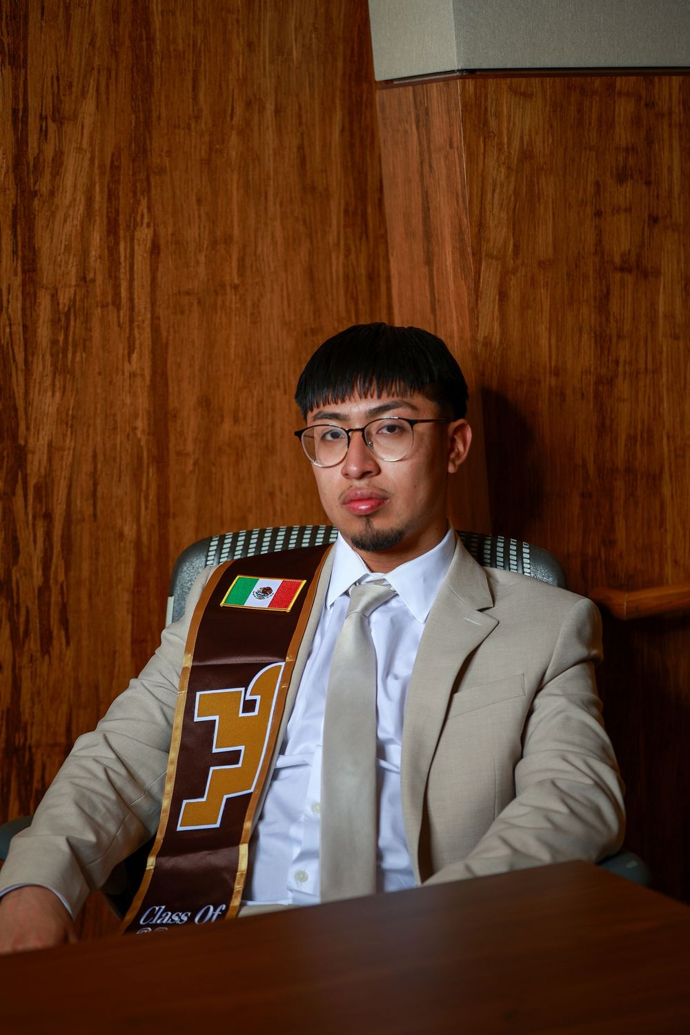 Graduate in tan suit and stole sits at desk with graduation memorabilia.
