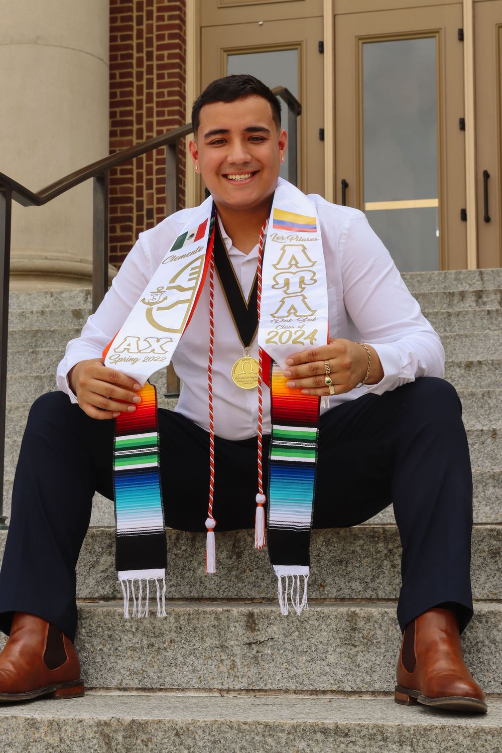Graduate wearing decorative sash with 2024 sits on stone steps near brick building.