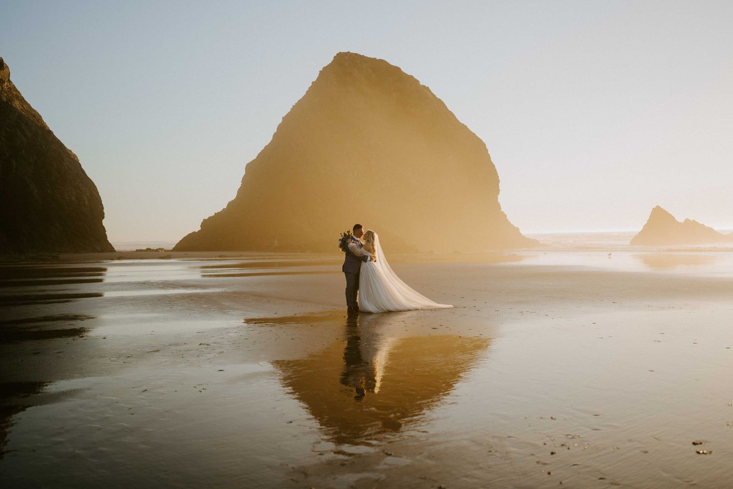 Wedding couple embracing on beach with dramatic sea stack rock formation at golden hour.