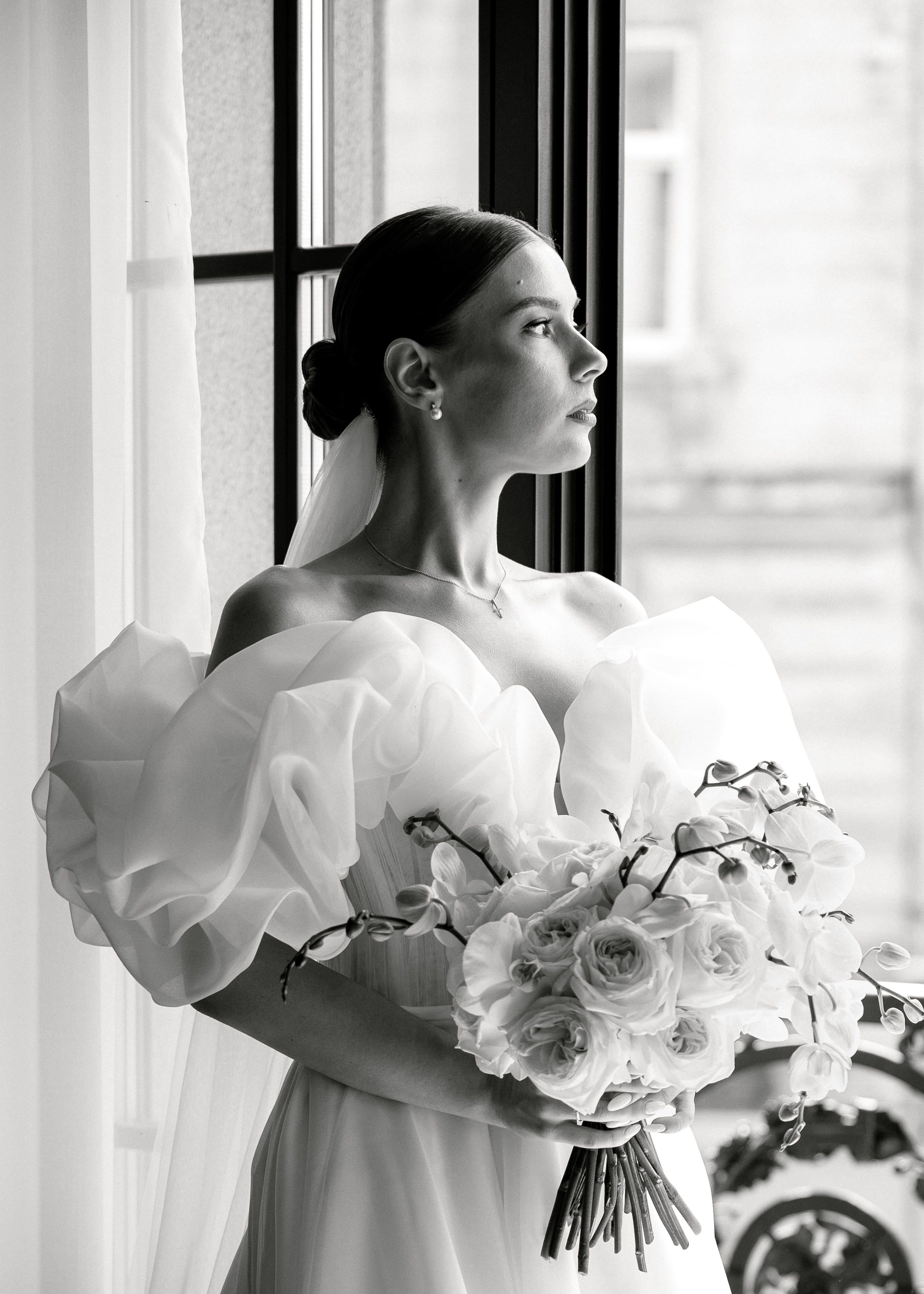 Black and white photo of a bride in a white dress with puff sleeves holding a bouquet of roses near a window.