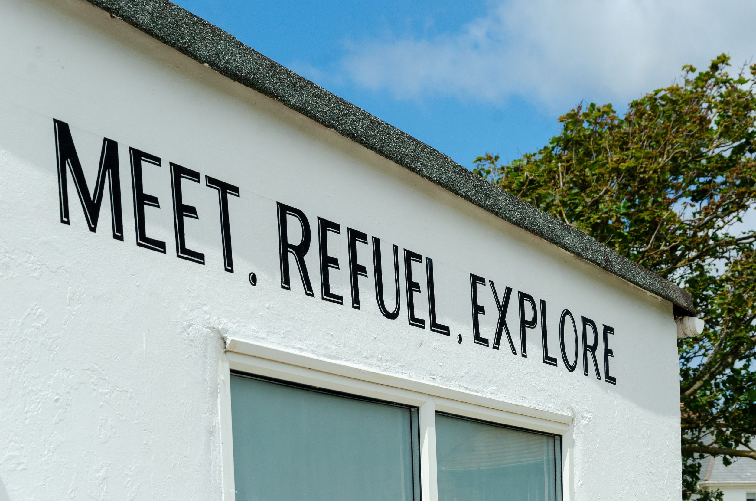 Sign on white building reads 'MEET, REFUEL, EXPLORE' against a blue sky with trees.