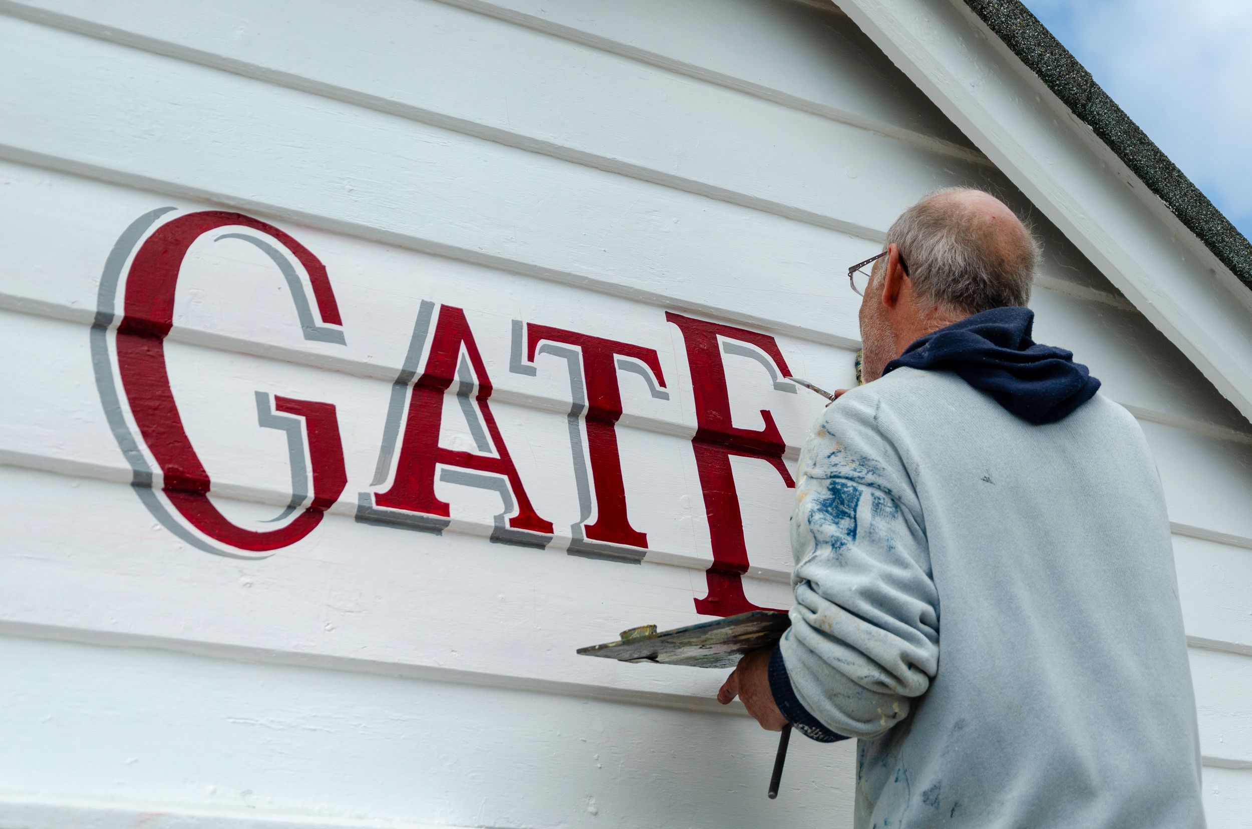 Person painting red letters spelling 'GATF' on white exterior building siding.