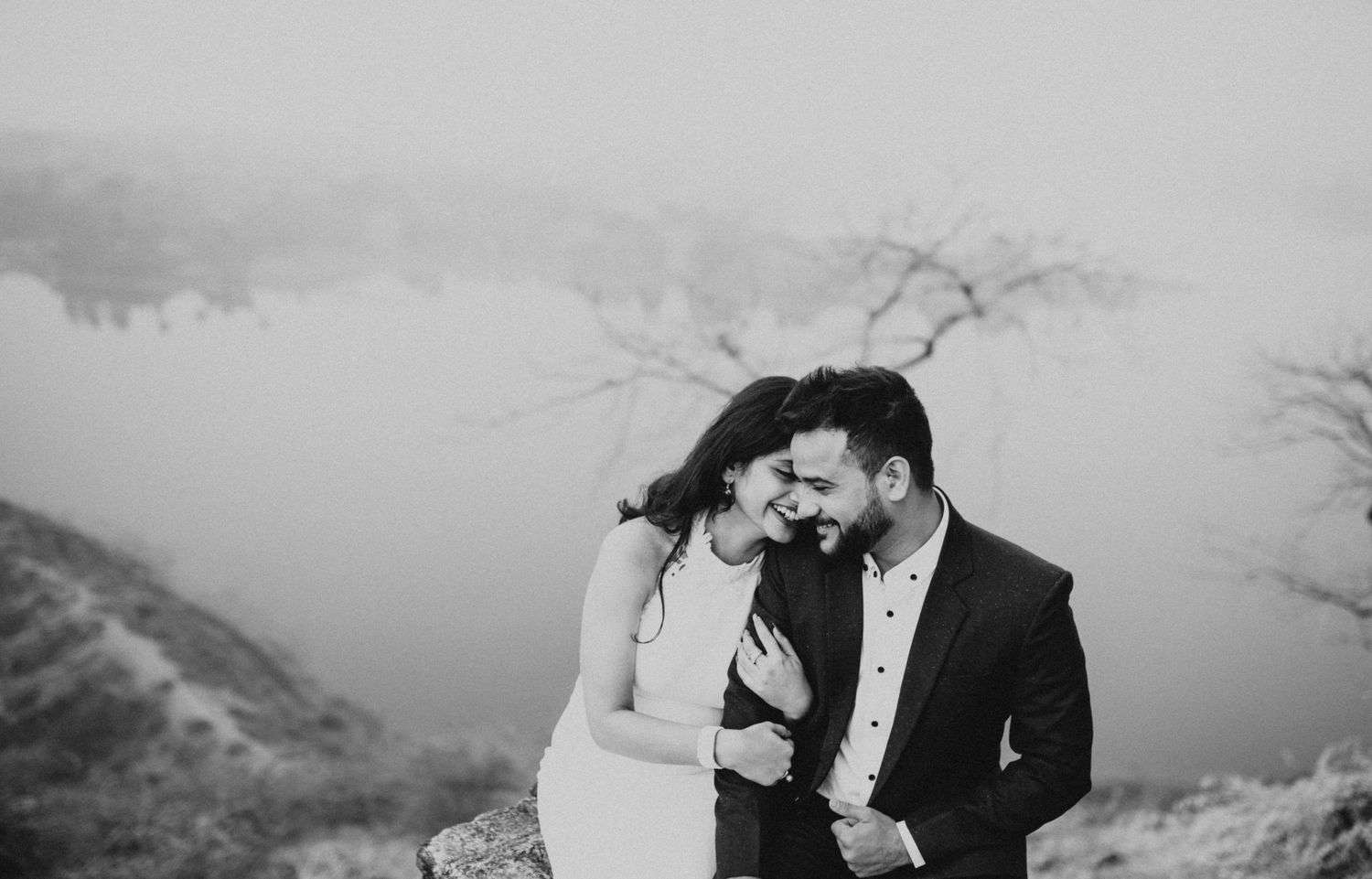 Black and white candid moment of affection captured against misty mountain backdrop.