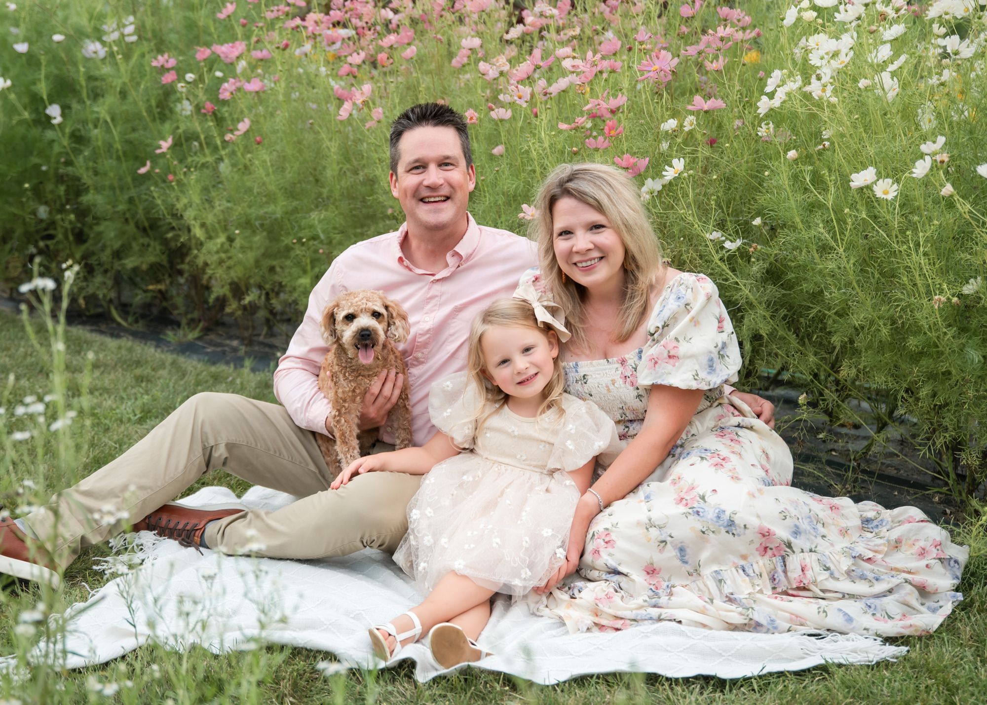 Family with small dog poses on blanket in garden with blooming cosmos flowers.