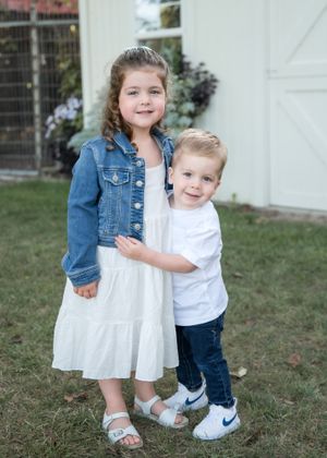 Two young siblings in coordinating outfits pose together on grass in front of a white building.