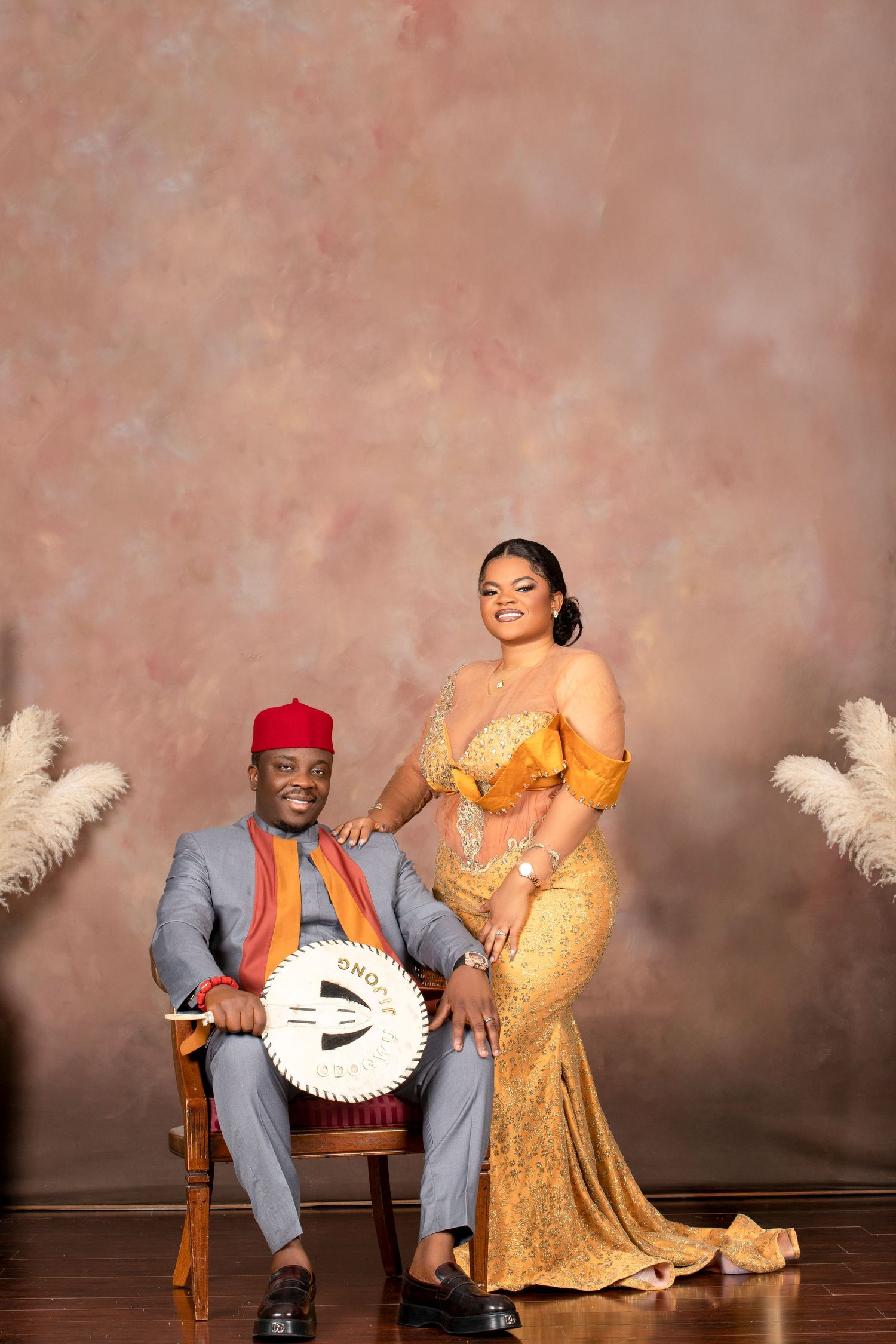 Formal portrait with traditional fan featuring gold gown and grey suit against neutral backdrop.