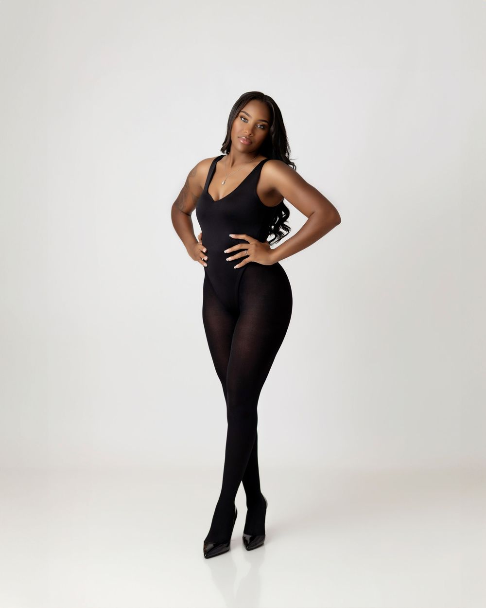 Woman in black bodysuit and opaque tights posing confidently with hands on hips against white background.