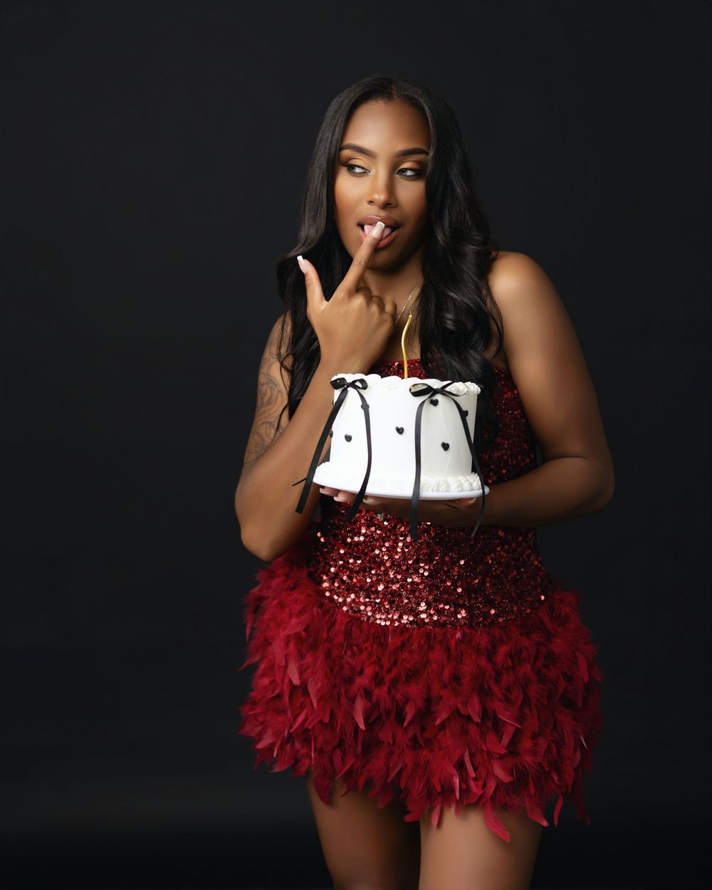 Woman in red sequin feather mini dress holding a decorated white cake with black bows in a studio setting.