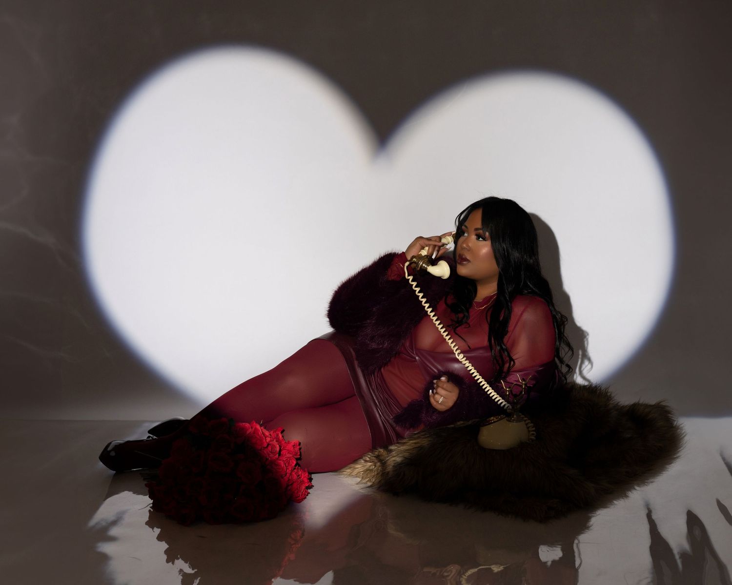Women in a burgundy dress poses on the floor with a heart-shaped light projection in the background.