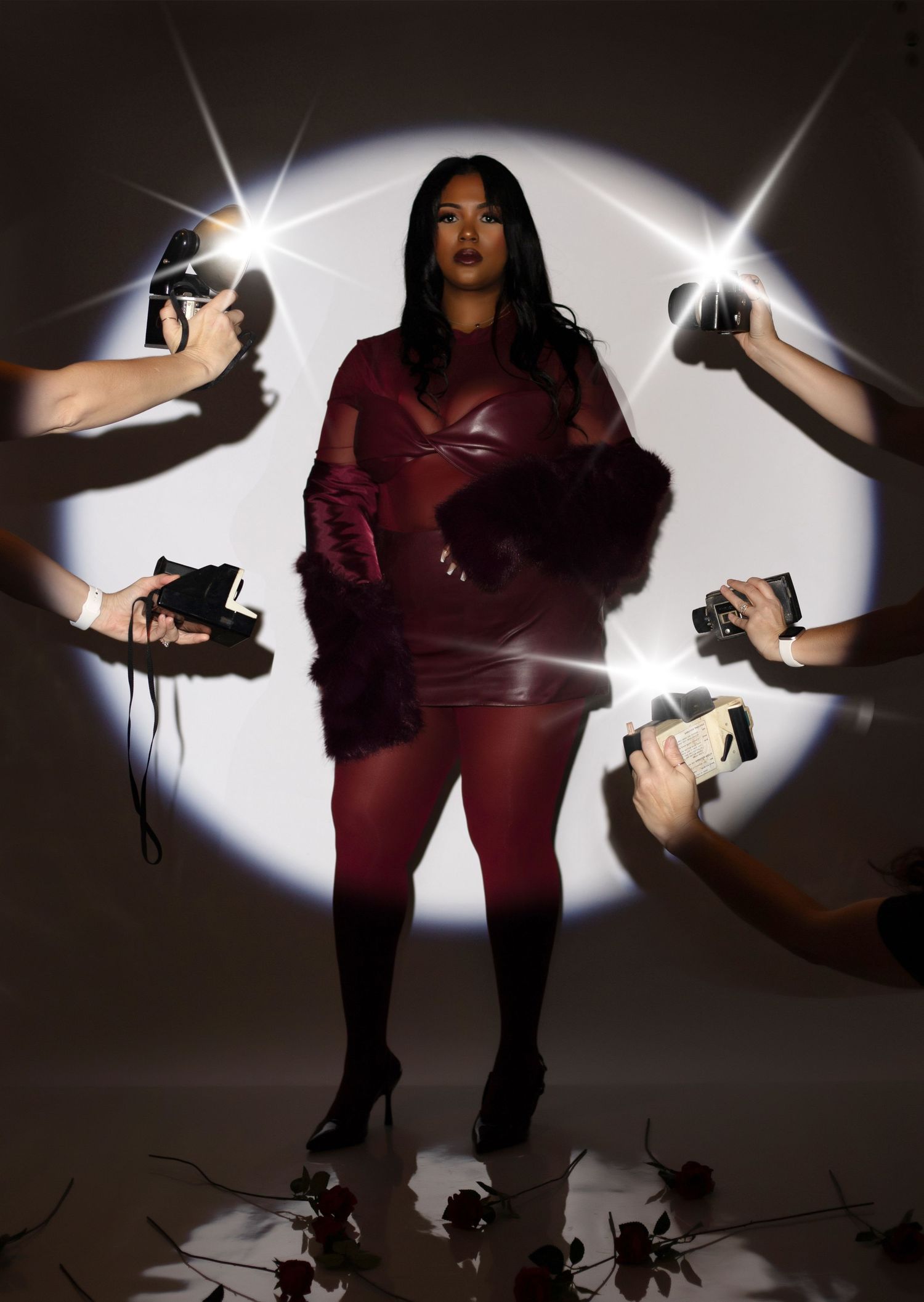 A women in burgundy miniskirt and fur coat stands in a spotlight while hands holding cameras reach in from the shadows.