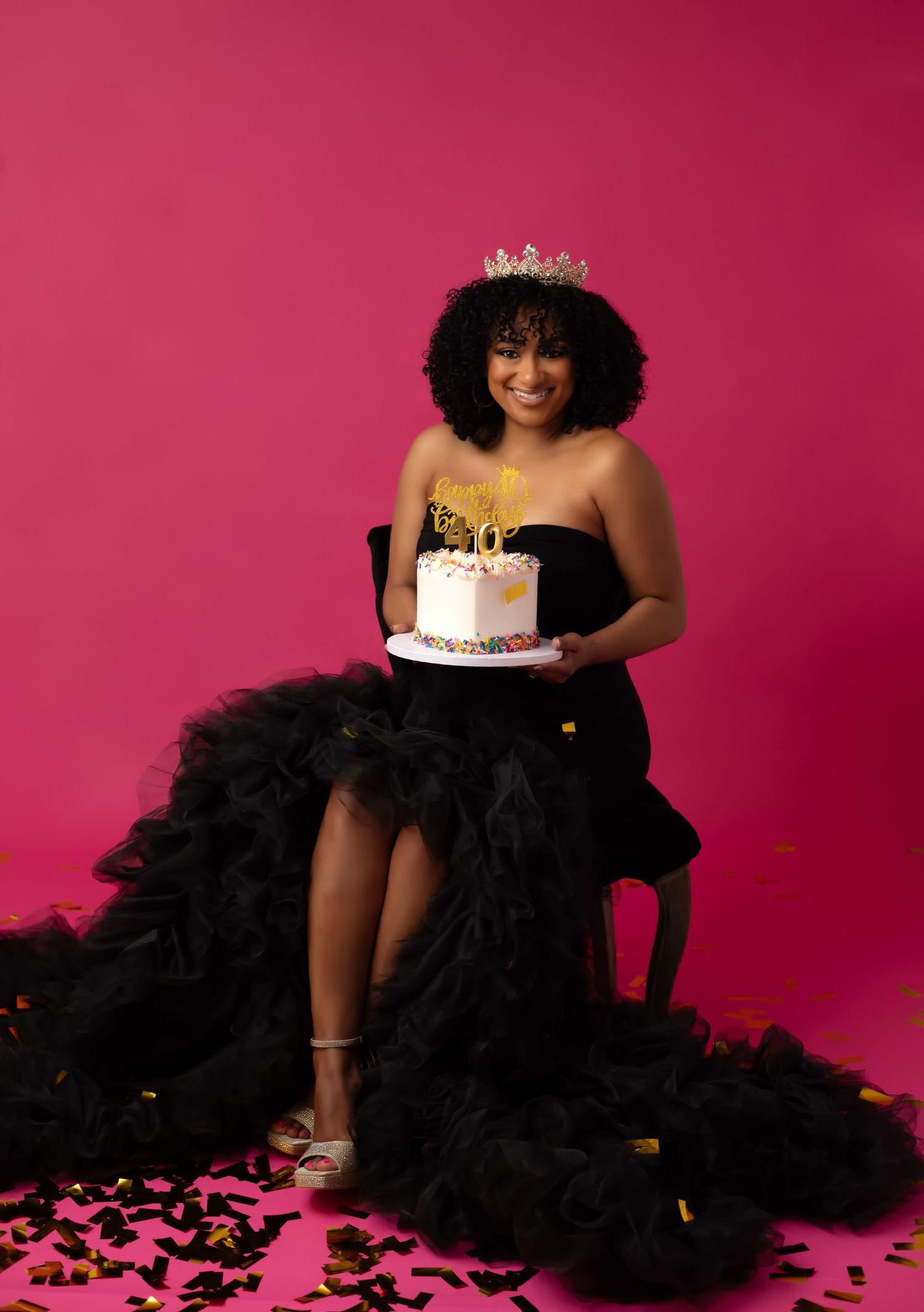 A glamorous Birthday photoshoot featuring a women in a black tulle gown against a vibrant pink backdrop with birthday cake and confetti.