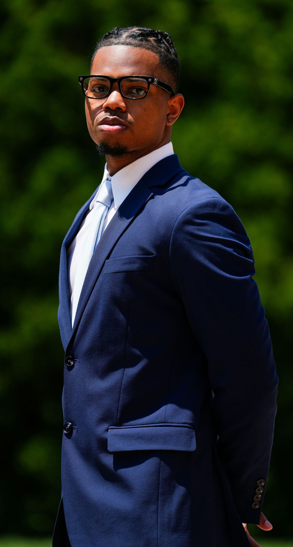 Professional portrait of someone in a fitted navy suit with glasses against a green background.