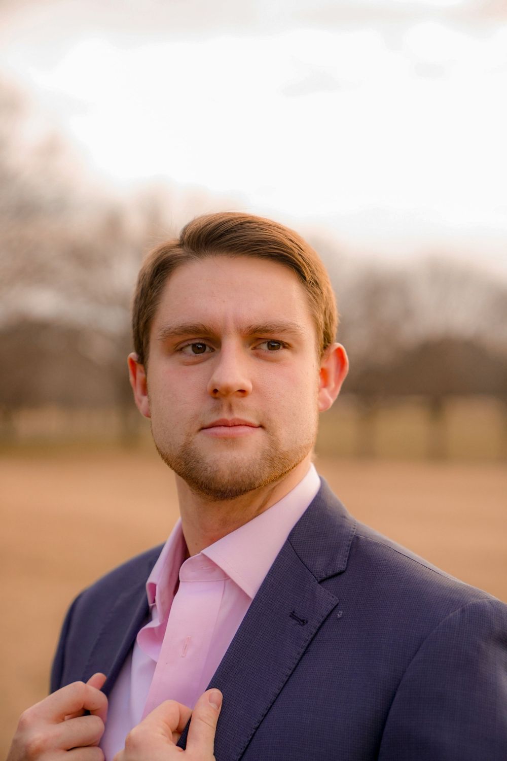 Professional headshot of someone in a navy suit and pink dress shirt at sunset.