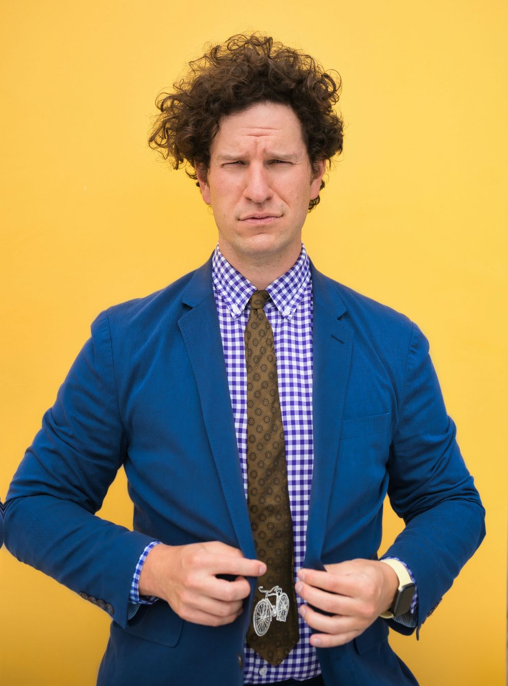Portrait of a person in a blue blazer and checkered shirt against a bright yellow background.
