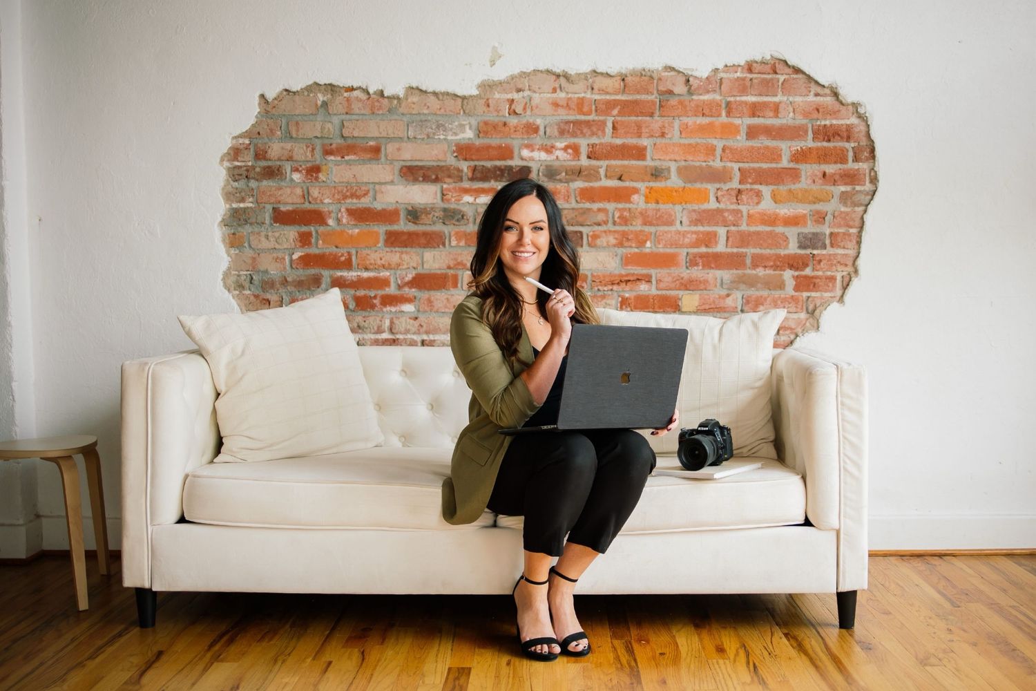 Woman sitting on white sofa with laptop, smiling in front of exposed brick wall on hardwood floors.