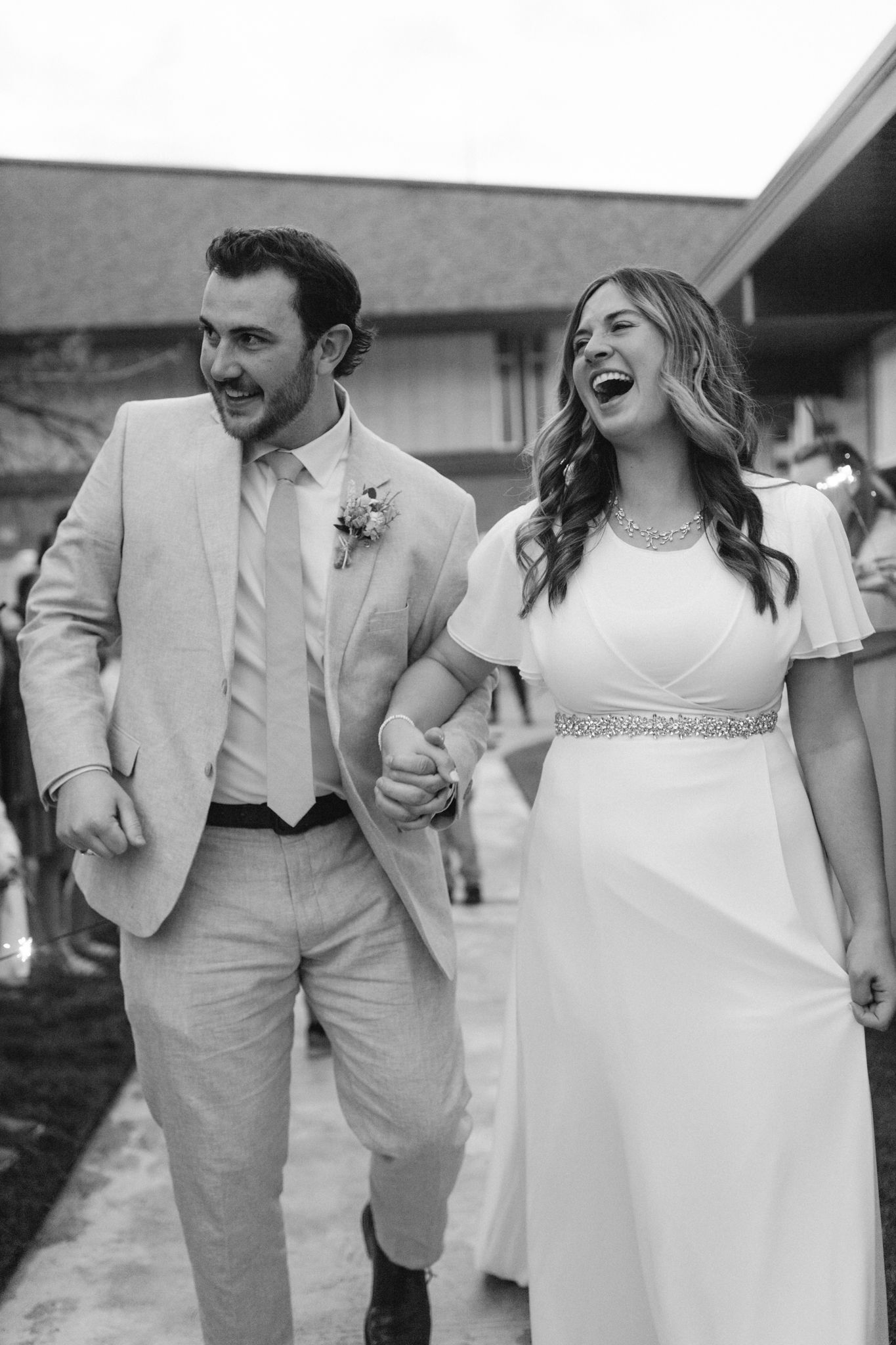 Laughing bride and groom walk hand in hand in black and white wedding photo outdoors.