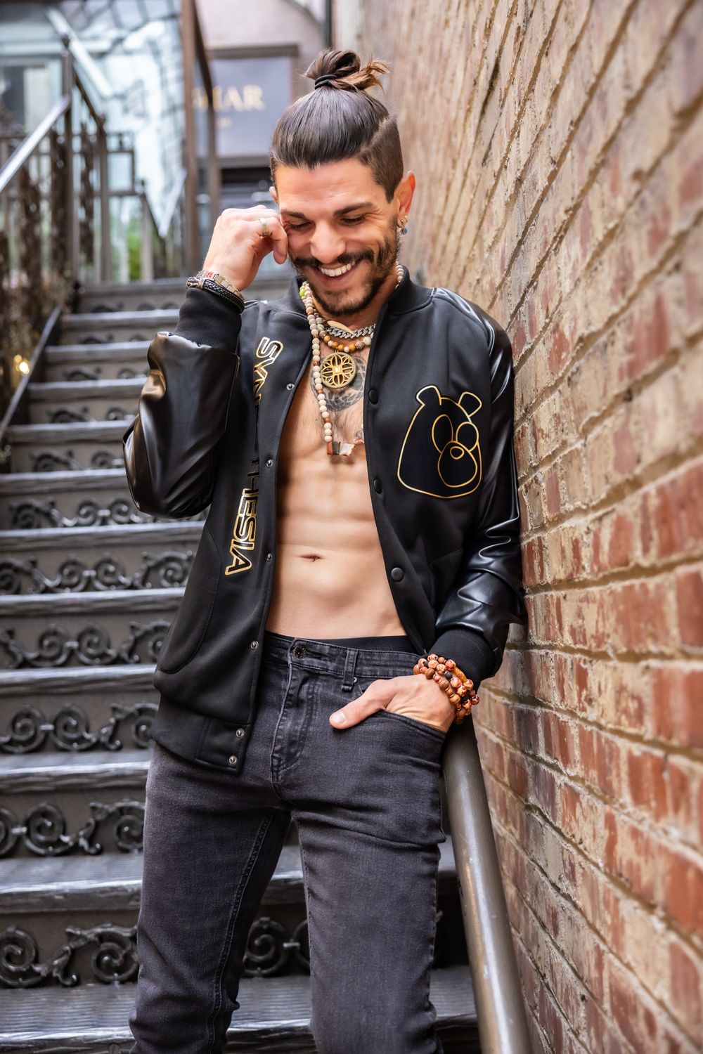 Urban portrait of man in leather jacket showing tattoos on stairs with brick wall background.