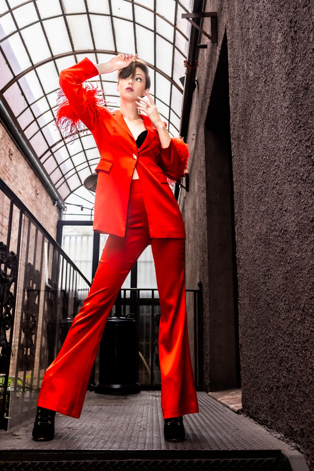 Fashion model in bright red power suit with feather details poses in industrial setting.