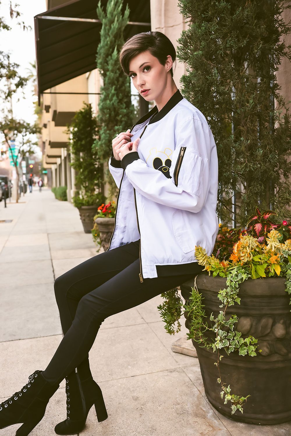 Model in white jacket and black boots poses on sidewalk next to decorative planter.