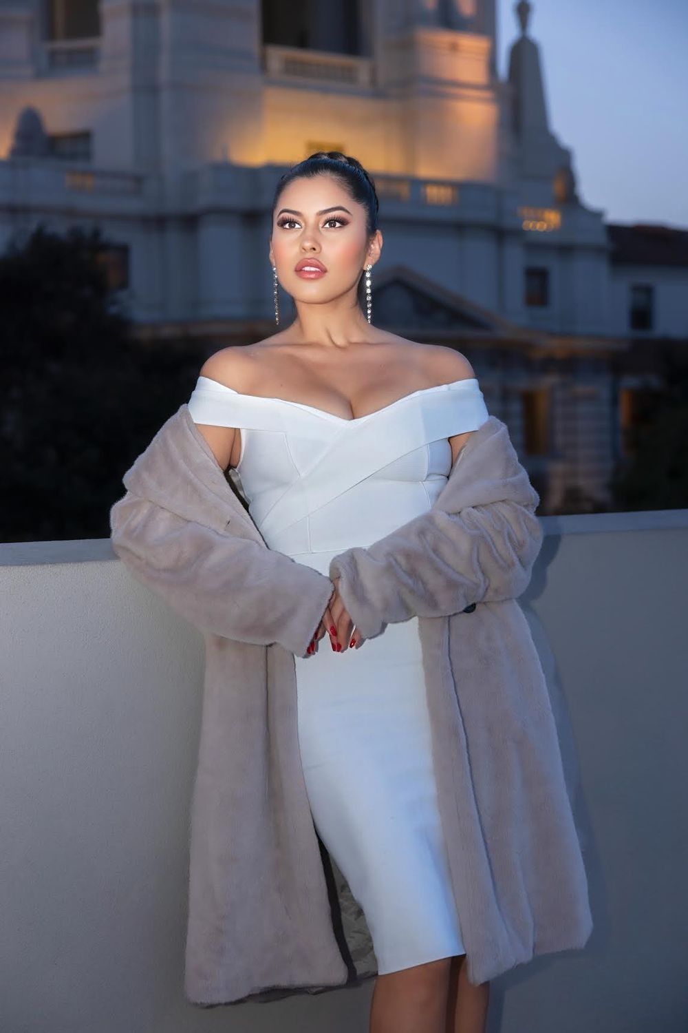 Evening portrait in white dress and fur coat on balcony with illuminated building backdrop.