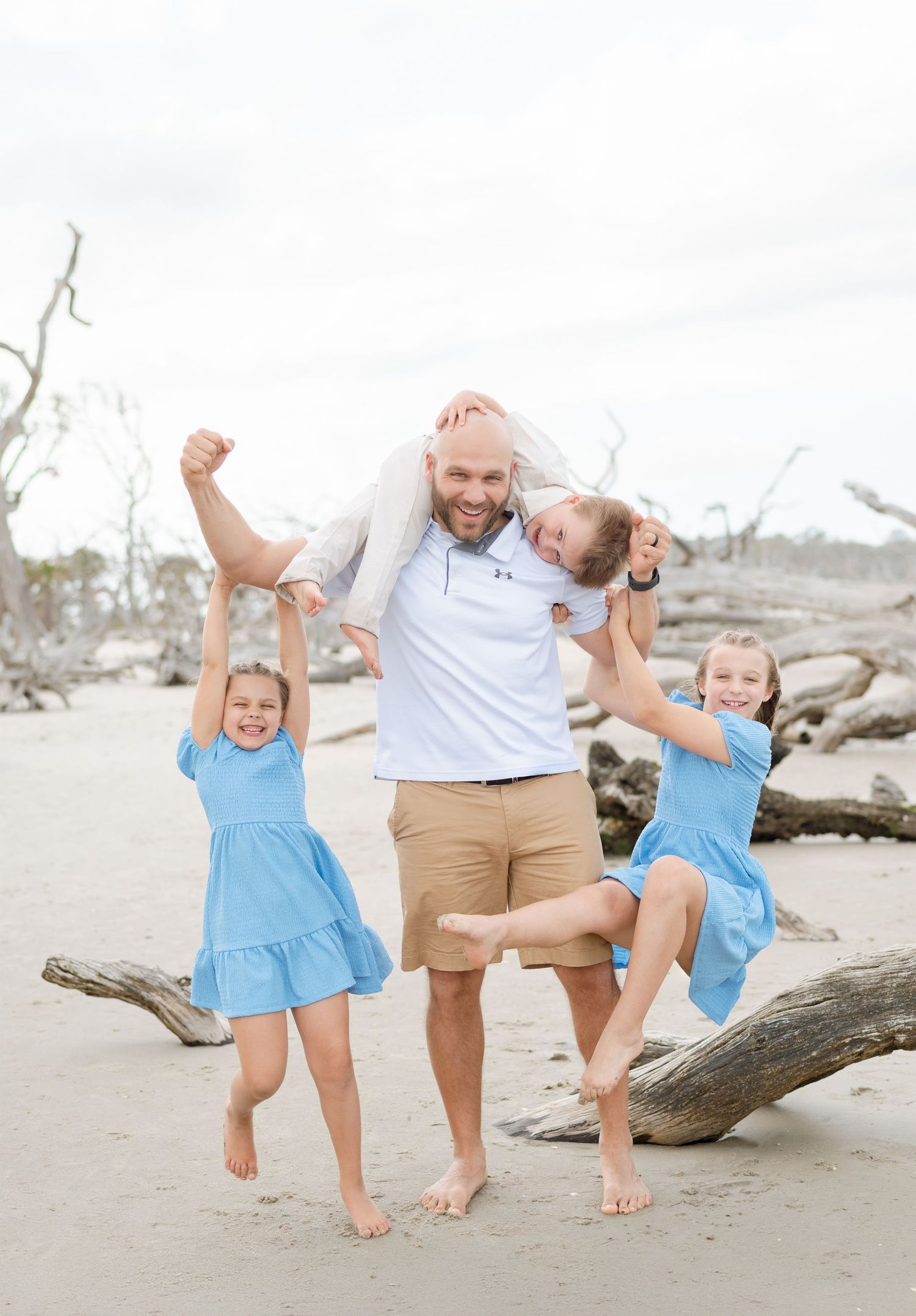 Father carrying a toddler on his shoulders while two girls in blue dresses jump beside him during a beach family portrait session.