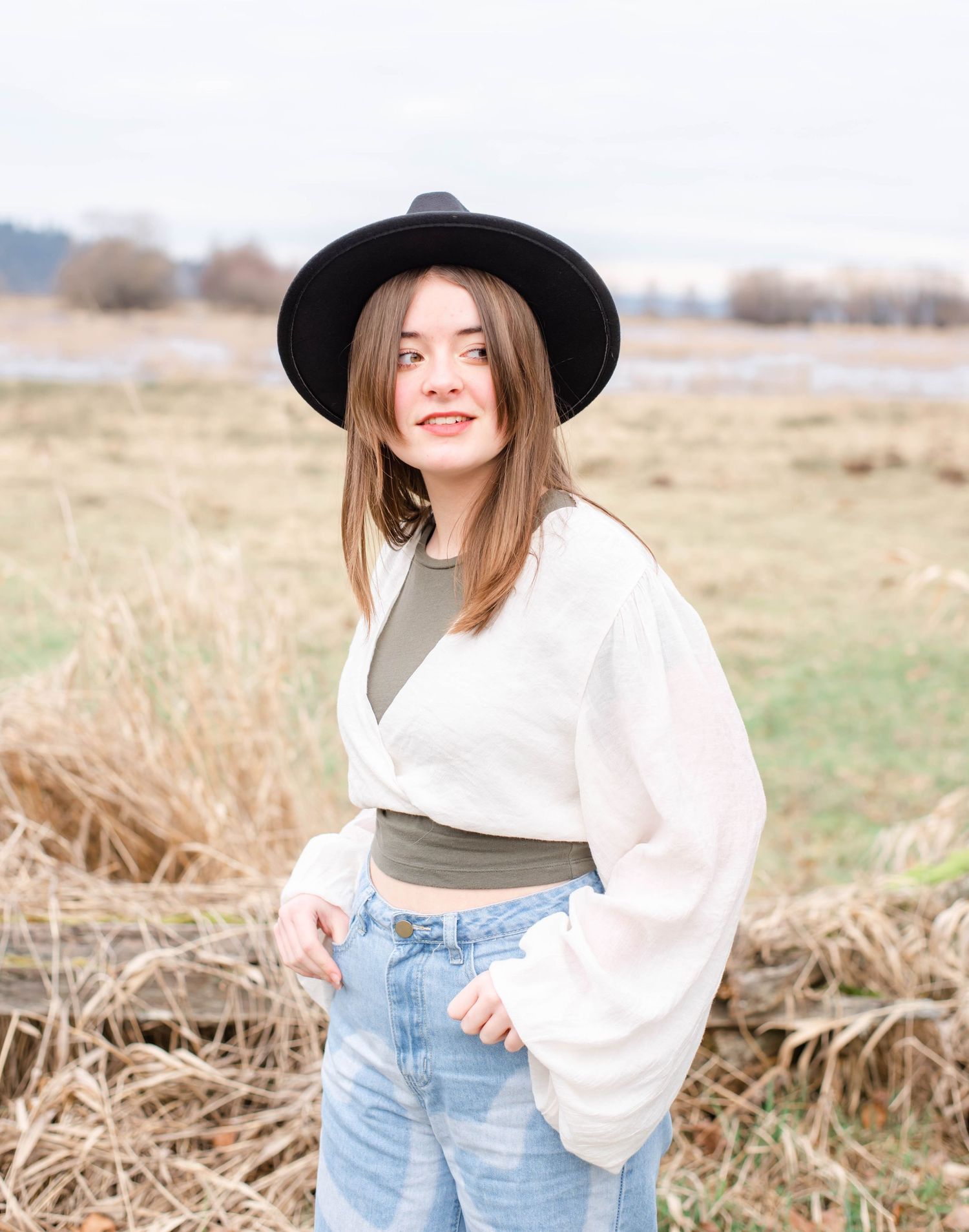 Wide-brim black hat and white blouse styled with denim in rustic field setting.