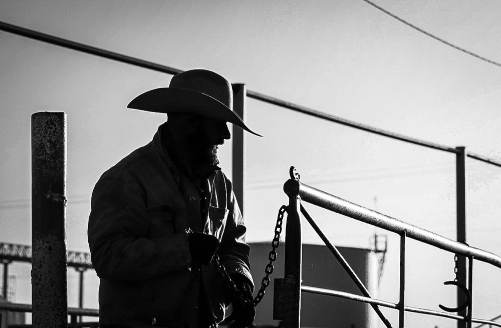 Black and white photograph of a silhouetted figure in a cowboy hat opening a gate against a bright sky backdrop.