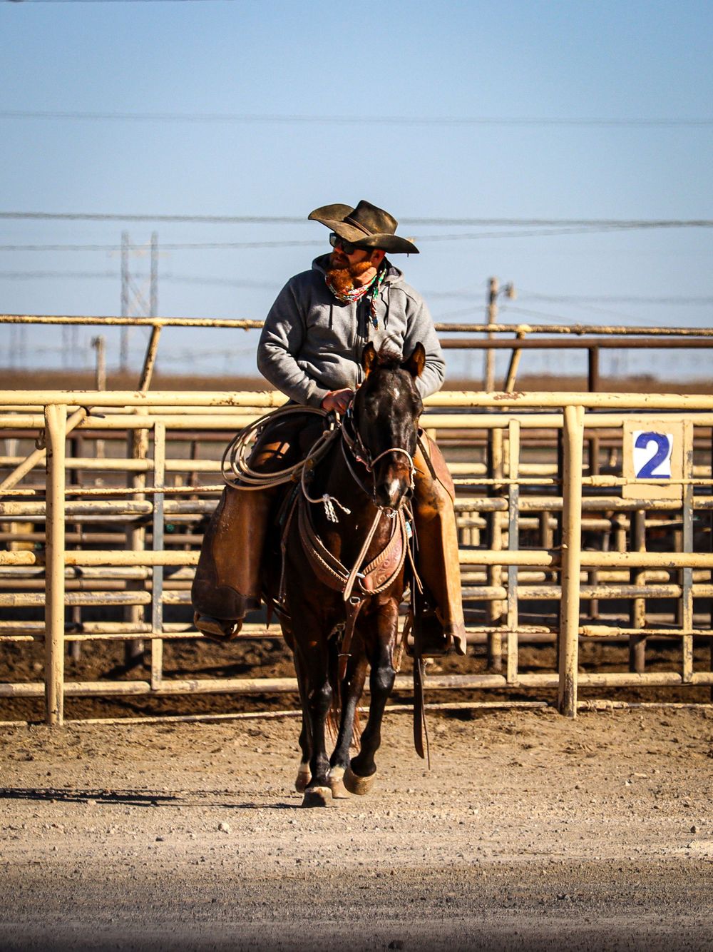 A cowboy rides a dark horse in a dusty corral with metal fencing and gates visible in the background.