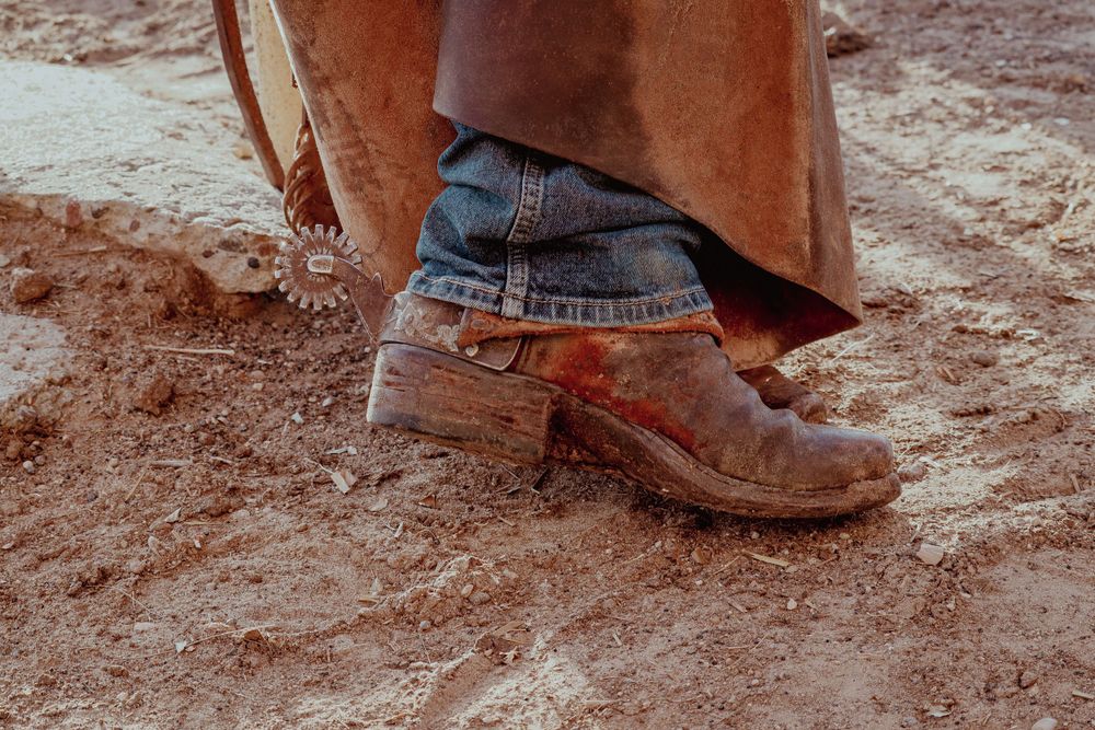 Close-up of a worn brown leather boot with spurs and leggins and denim jeans standing on dusty red soil.