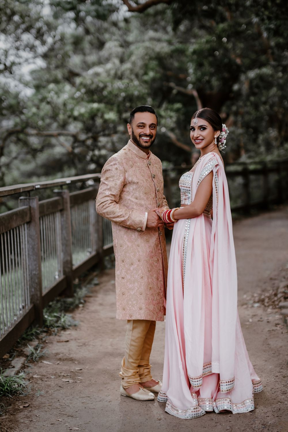 Couple in traditional Indian wedding attire posing on wooden bridge pathway.
