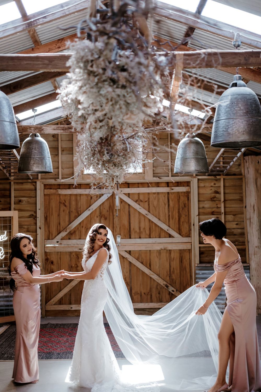 Bridesmaids helping adjust bride's veil in rustic barn setting with hanging lights.