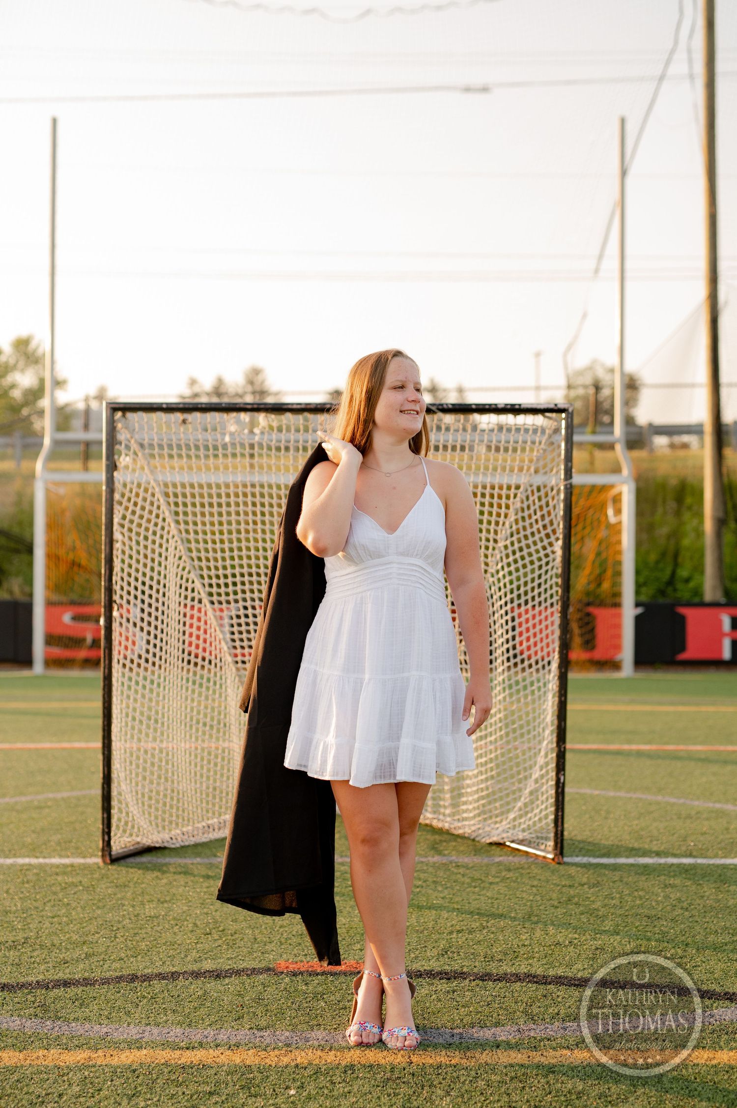Graduate in white sundress and black graduation gown poses against lacrosse goal on sports field.