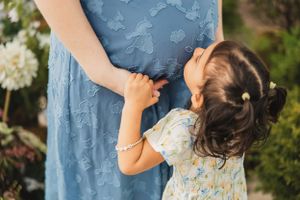 A child kisses her mother's pregnant belly while holding her hands surrounded by florals in a greenhouse.